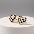 A pair of heart-shaped stud earrings with a black and white checkerboard pattern and gold-tone borders, displayed on a white cylindrical pedestal against a gray background.