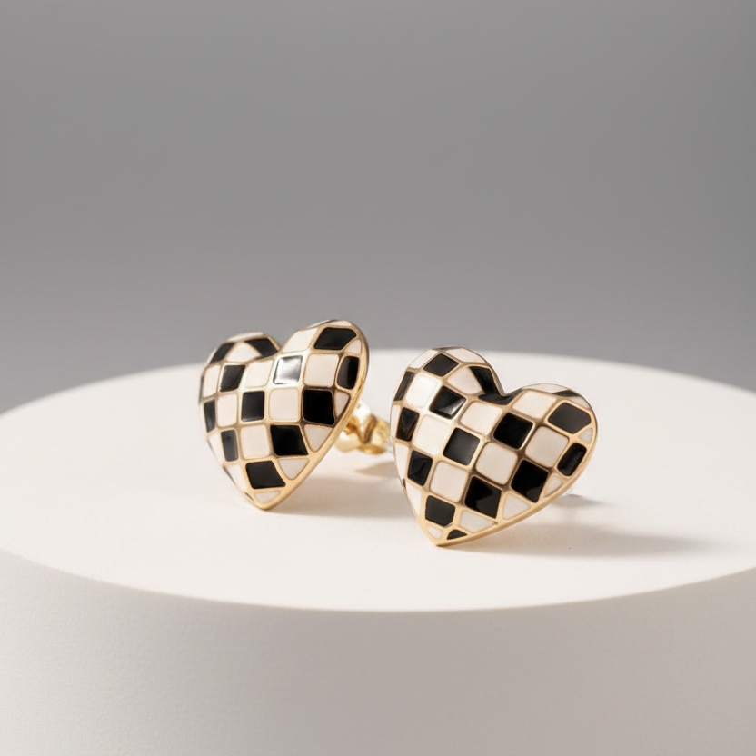 A pair of heart-shaped stud earrings with a black and white checkerboard pattern and gold-tone borders, displayed on a white cylindrical pedestal against a gray background.