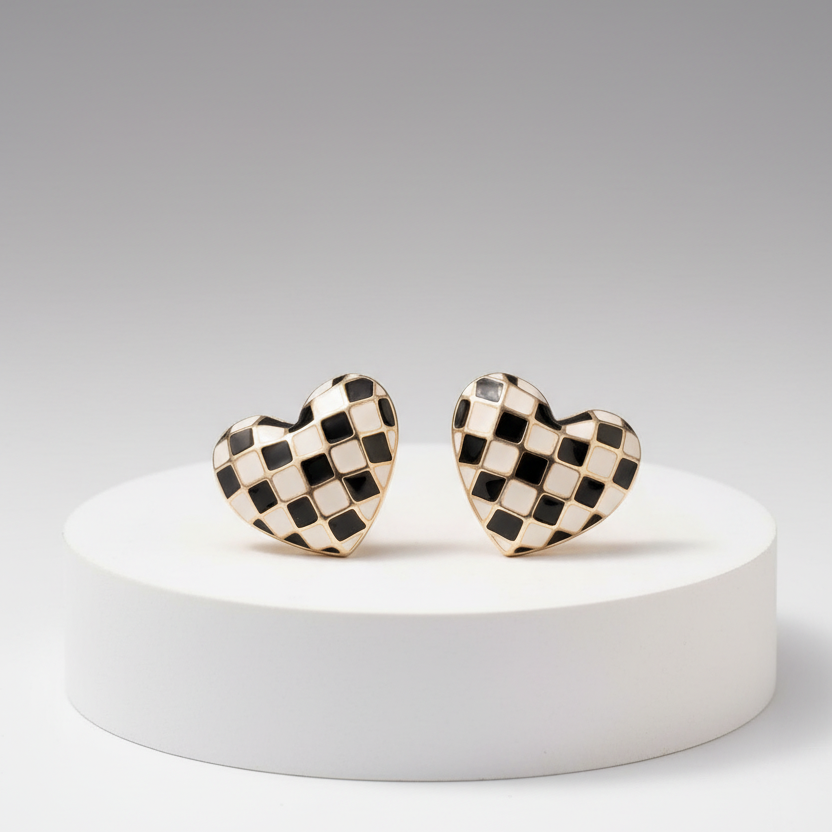 A pair of heart-shaped stud earrings with a black and white checkerboard pattern and gold-tone edges, displayed on a white cylindrical pedestal against a gray gradient background.