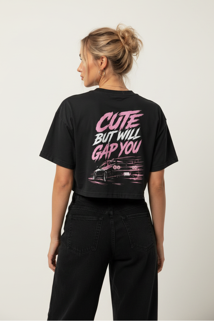 A woman shown from the back wearing a black cropped graphic T-shirt with pink and white text reading 'CUTE BUT WILL GAP YOU' and a car illustration, paired with black pants, against a plain light background.