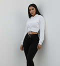 A woman modeling a white cropped hoodie with front lace-up detailing and black skinny jeans, standing against a plain light gray wall.