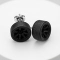 A pair of black stud earrings designed to look like miniature car wheels with textured treads and multi-spoke rims, shown on a white circular pedestal.