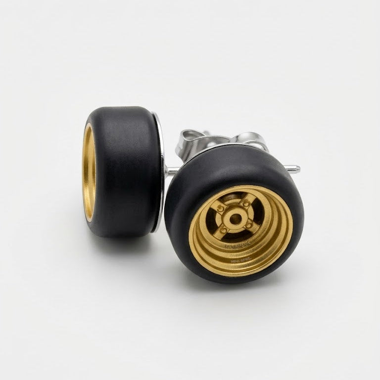 A pair of stud earrings shaped like miniature car wheels, featuring a matte black outer rim and a polished gold-toned center hub, shown on a clean white background.