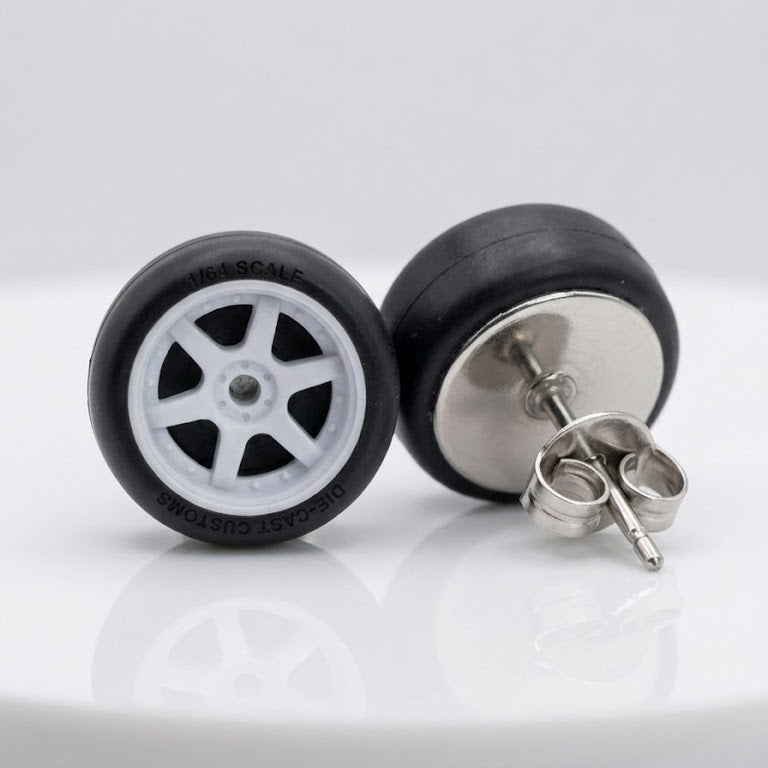 A pair of stud earrings designed to look like miniature toy car wheels, featuring black tires and white multi-spoke rims, with one earring turned to show the silver butterfly back.