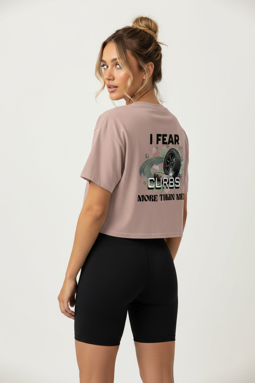 A woman shown from the back wearing a cropped dusty pink t-shirt with a graphic on the back that reads 'I FEAR CURBS MORE THAN ME' and black fitted biker shorts, against a plain light gray studio background.