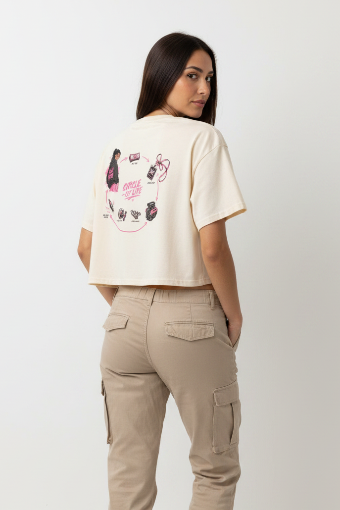 A woman shown from the back wearing a cream-colored short-sleeve crop top with a colorful graphic print on the back, paired with beige cargo pants, against a plain white studio background.