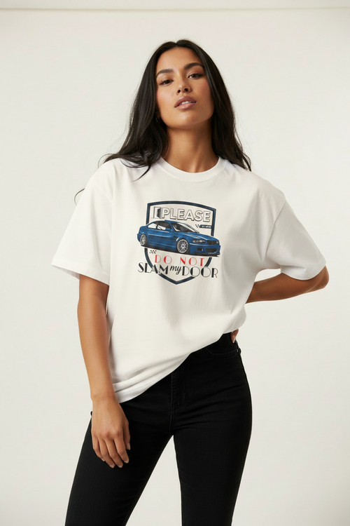 A woman modeling a white graphic t-shirt with a blue car and 'PLEASE DO NOT SLAM MY DOOR' print, paired with black pants, against a plain light background.