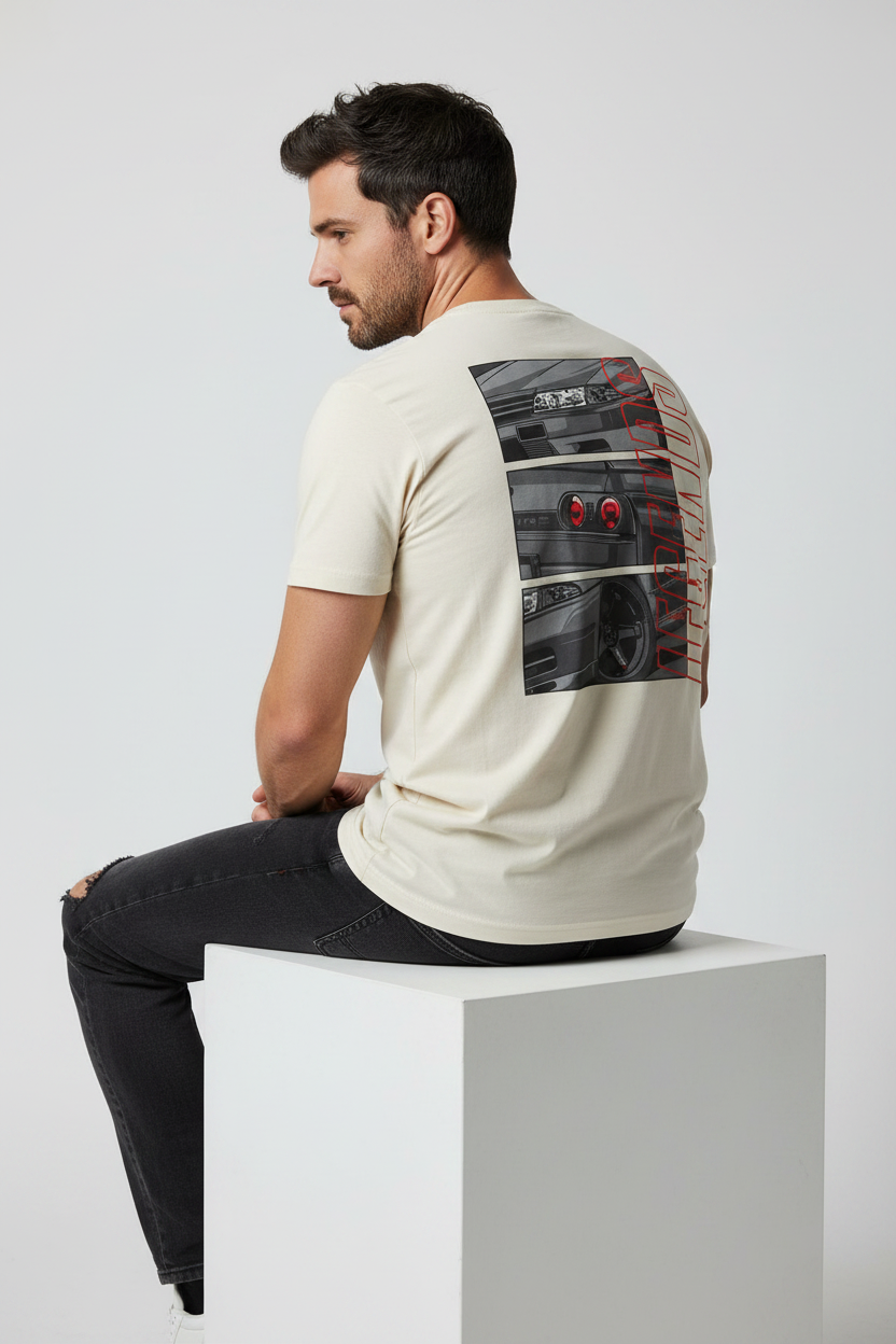 A man wearing a cream-colored t-shirt with a large graphic print of a black sports car on the back, sitting on a white block against a light gray background.