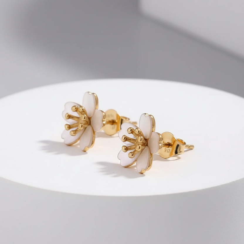 A pair of gold-tone stud earrings with white enamel flower petals and gold stamens, displayed on a white cylindrical pedestal against a light gray background.