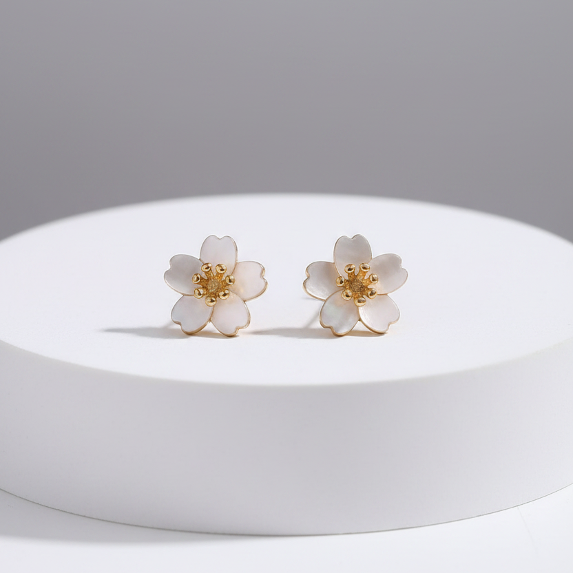A pair of small flower-shaped stud earrings with white petals and gold centers, displayed on a white round pedestal against a soft gray background.