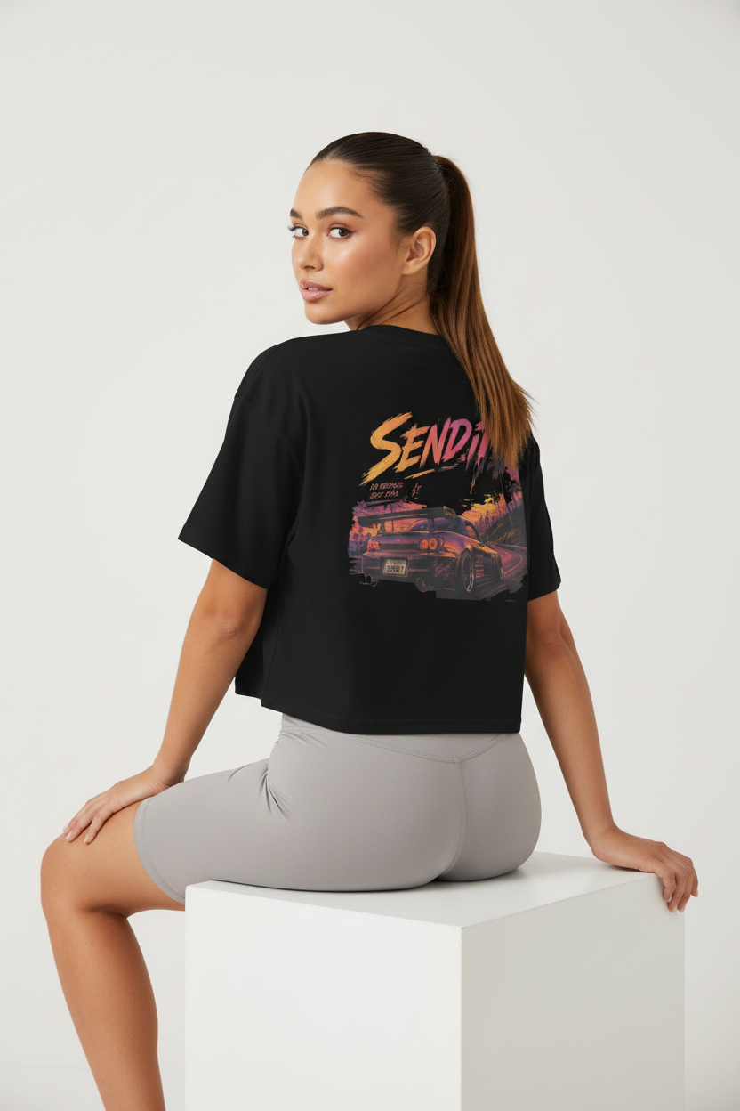 A woman wearing a black cropped t-shirt with a colorful 'Send It' graphic on the back, paired with light gray fitted shorts, sitting on a white display block against a light gray studio background.